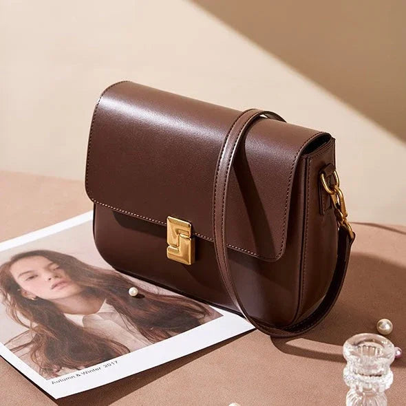 Brown leather crossbody bag with gold clasp on beige table, magazine and pearls nearby