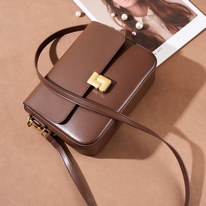 Brown leather crossbody bag with gold buckle on tan surface, fashion magazine and pearls