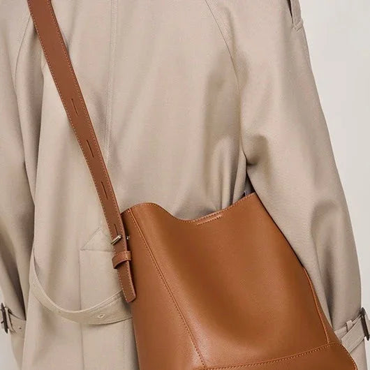 Tan leather bucket bag with adjustable strap worn over a beige trench coat