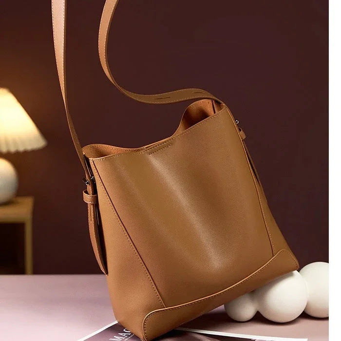Brown leather bucket bag with long strap on table, modern minimalist setting