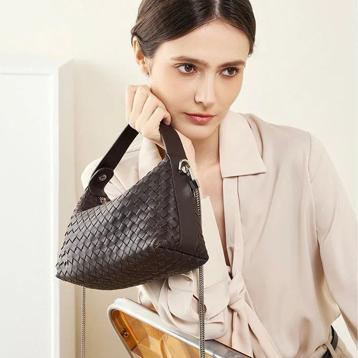 Woman holding a dark brown woven leather handbag, wearing a light beige blouse indoors