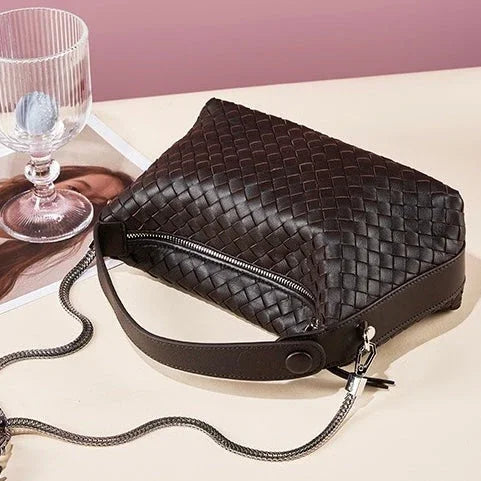 Woven brown leather crossbody bag with zipper on a beige table, glass cup in background
