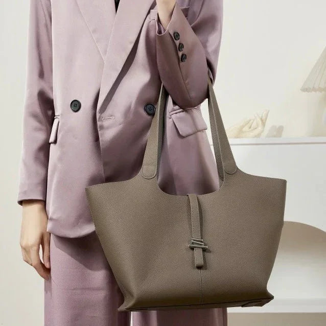 Woman in mauve suit holding large taupe leather tote bag in minimalist indoor setting