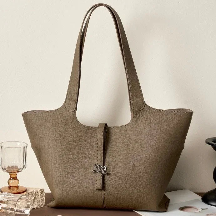 Taupe leather tote bag with double handles and clasp detail on neutral tabletop setting