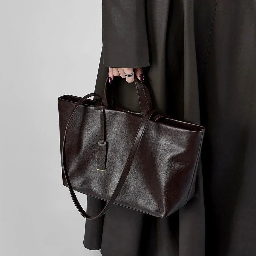 Hand holding a dark brown leather tote bag with tag, black coat in background