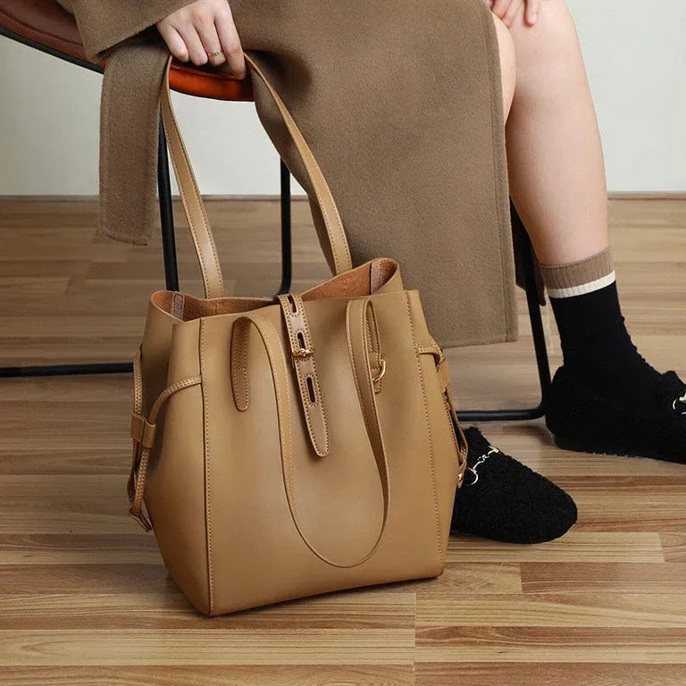 Tan leather tote bag on wood floor beside seated person in brown coat and black shoes