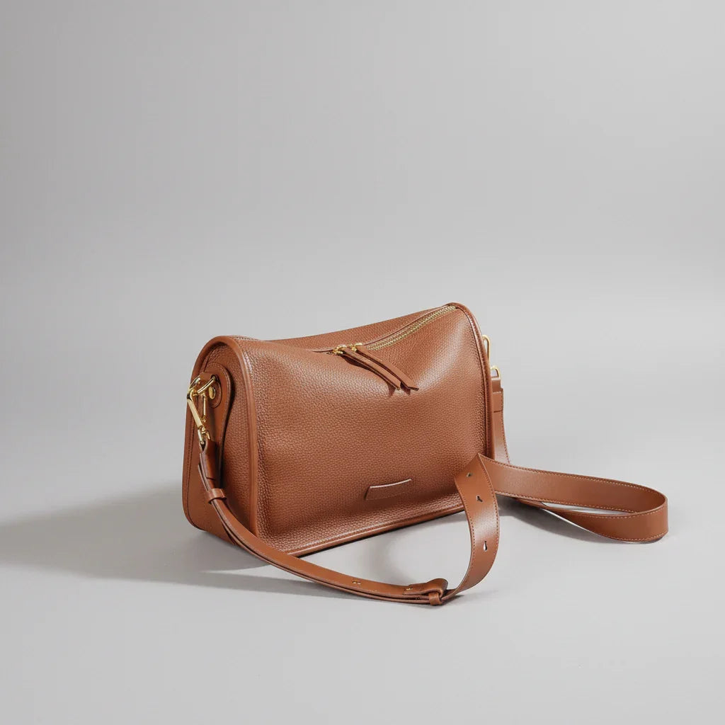 Brown leather crossbody bag with gold zipper on textured surface, rustic background