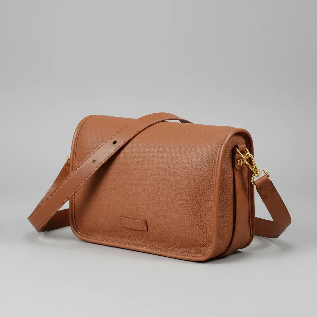 Brown leather crossbody bag on woven surface, with gold hardware and adjustable strap