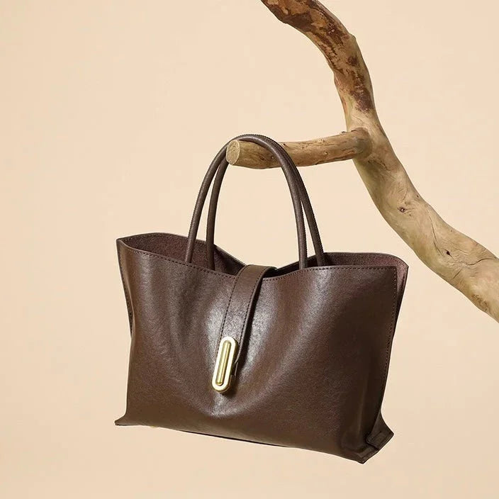 Brown leather tote bag with gold clasp, hanging on a tree branch, beige background