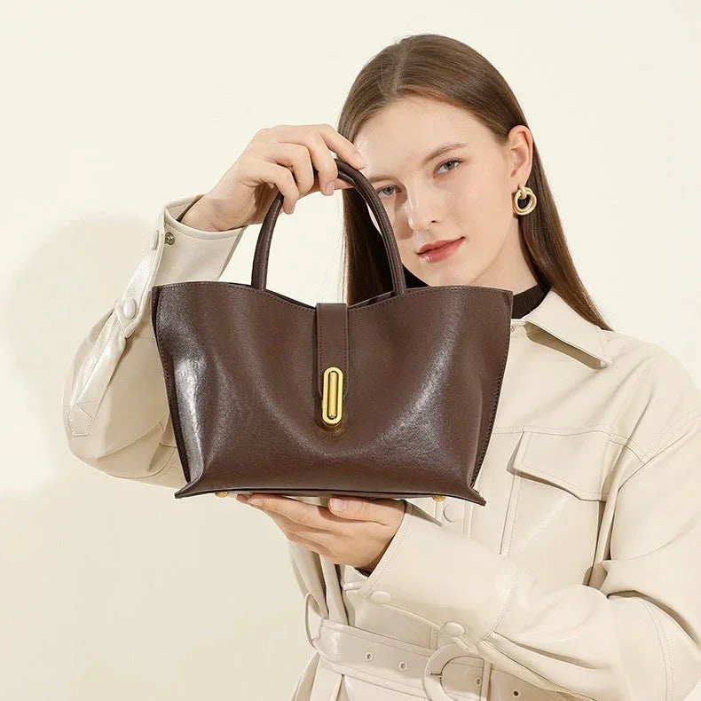 Woman holding brown leather handbag with gold accent, wearing cream trench coat