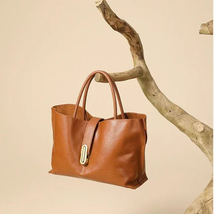 Brown leather tote handbag with gold accent hangs on a branch, beige background