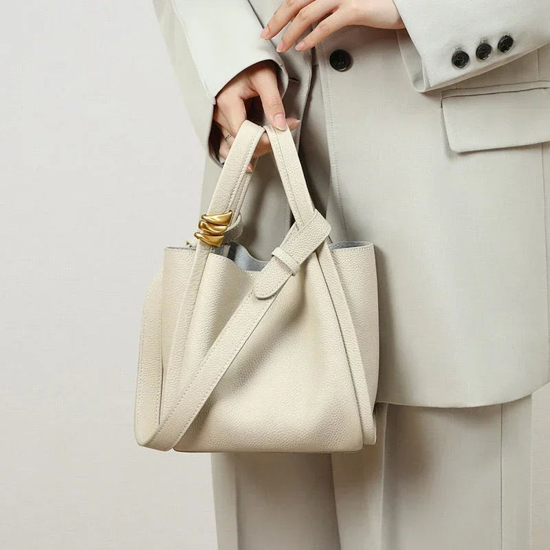 Woman in a cream suit holding a beige leather handbag with gold accent and shoulder strap