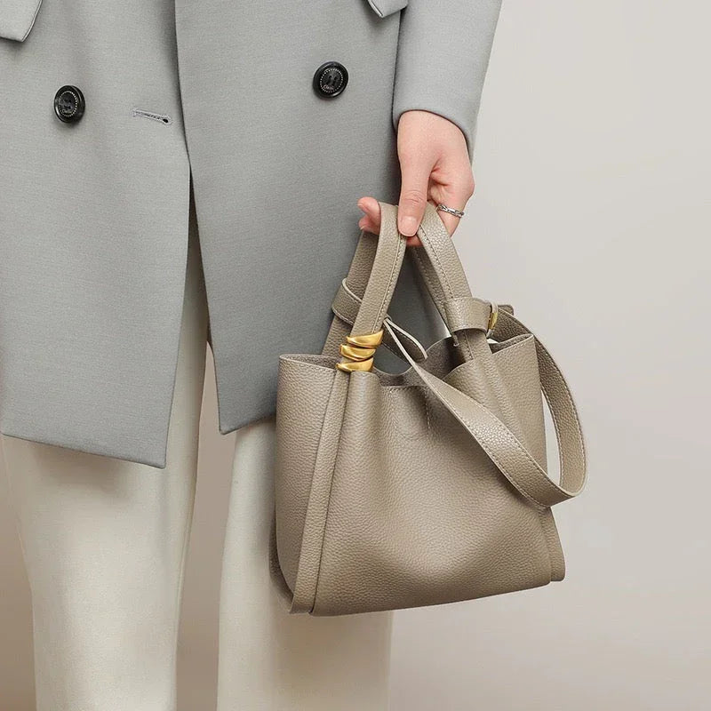 Woman holding taupe leather bucket bag, wearing light grey coat and white pants