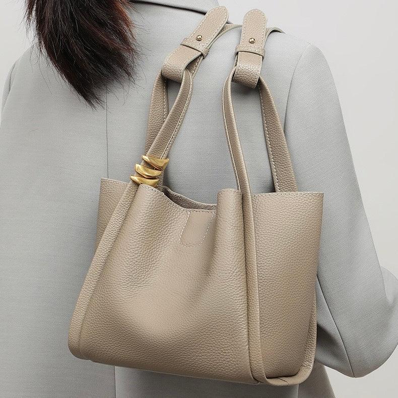 Taupe leather tote bag with gold rings on handle, worn over a grey blazer