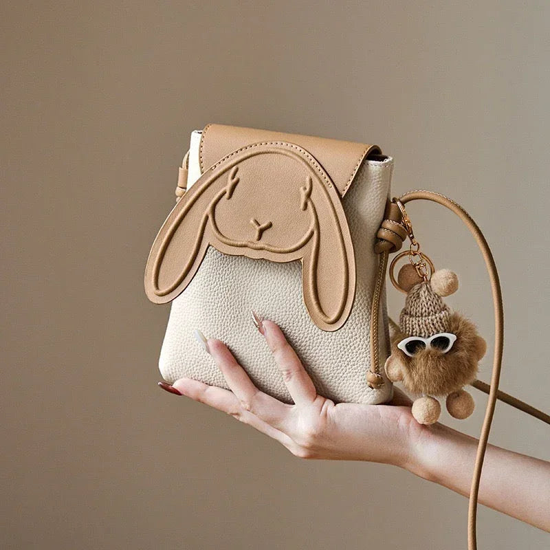 Beige crossbody bag with bunny face design and plush bear keychain, held by a hand