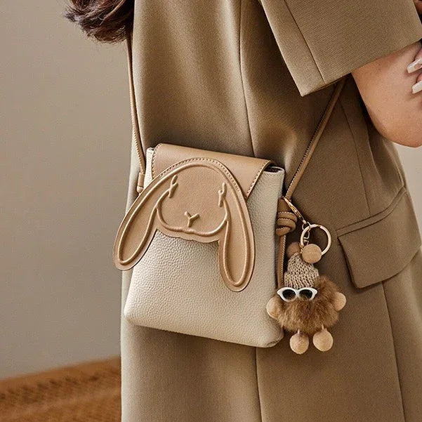 Beige crossbody bag with bunny face design and cute plush keychain, worn on shoulder.