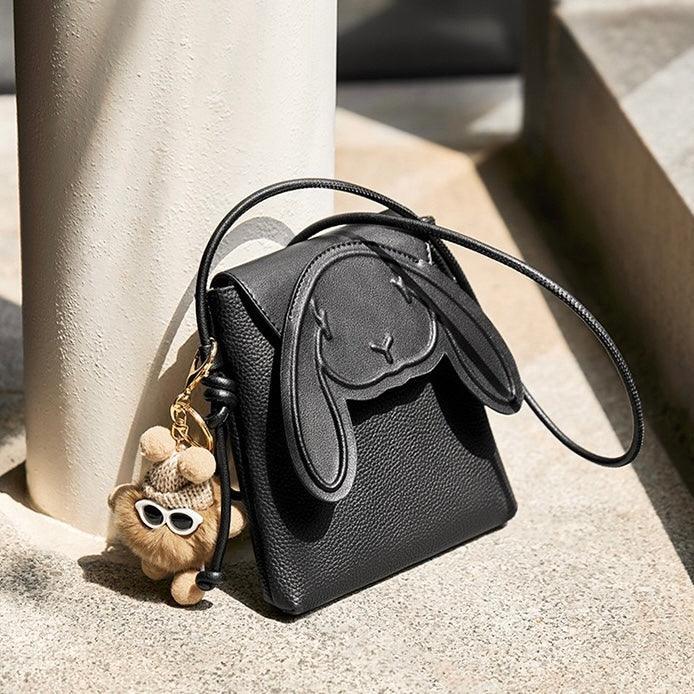 Black leather bunny ear crossbody bag with plush bear keychain on stone steps