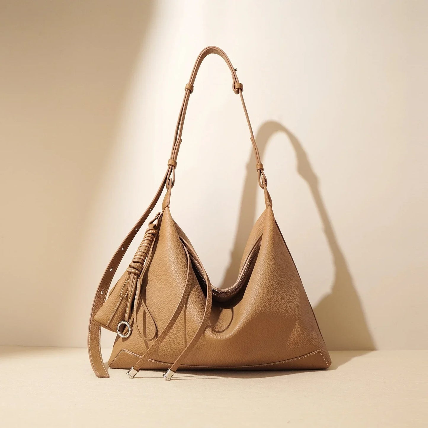Tan leather hobo bag with adjustable strap and decorative tassel on beige background