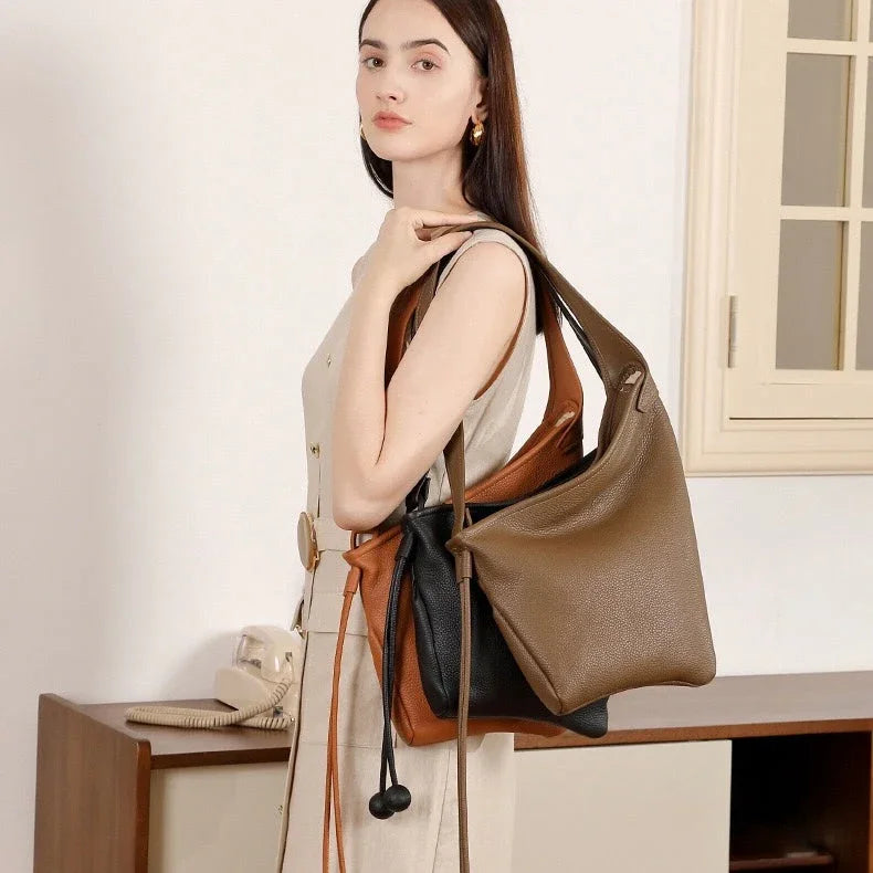 Woman in beige dress modeling three large leather tote bags in brown, tan, and black indoors