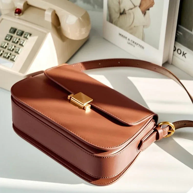 Brown leather crossbody bag with gold clasp on white desk near vintage phone and books