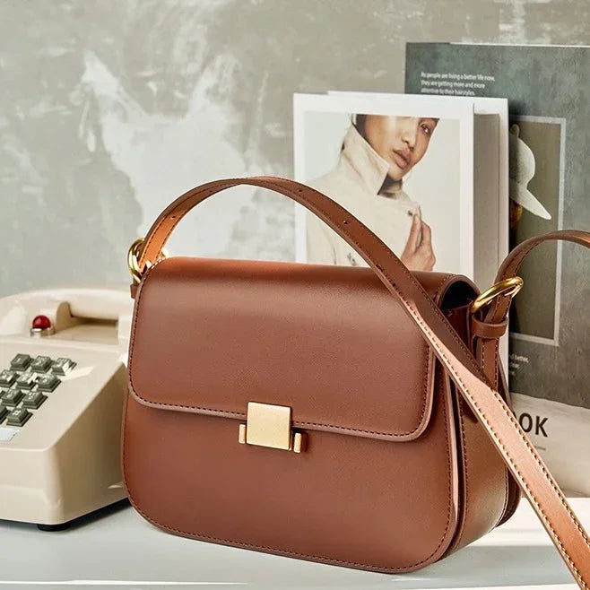Brown leather shoulder bag with gold clasp on desk beside vintage telephone and magazines