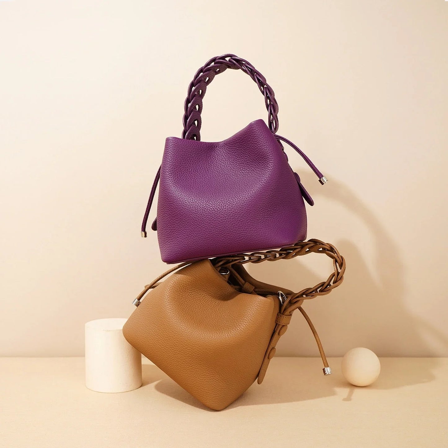 Two leather handbags, one purple and one tan, with braided handles on a beige background