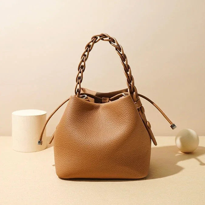 Tan leather bucket bag with braided handle on beige background, minimalist style