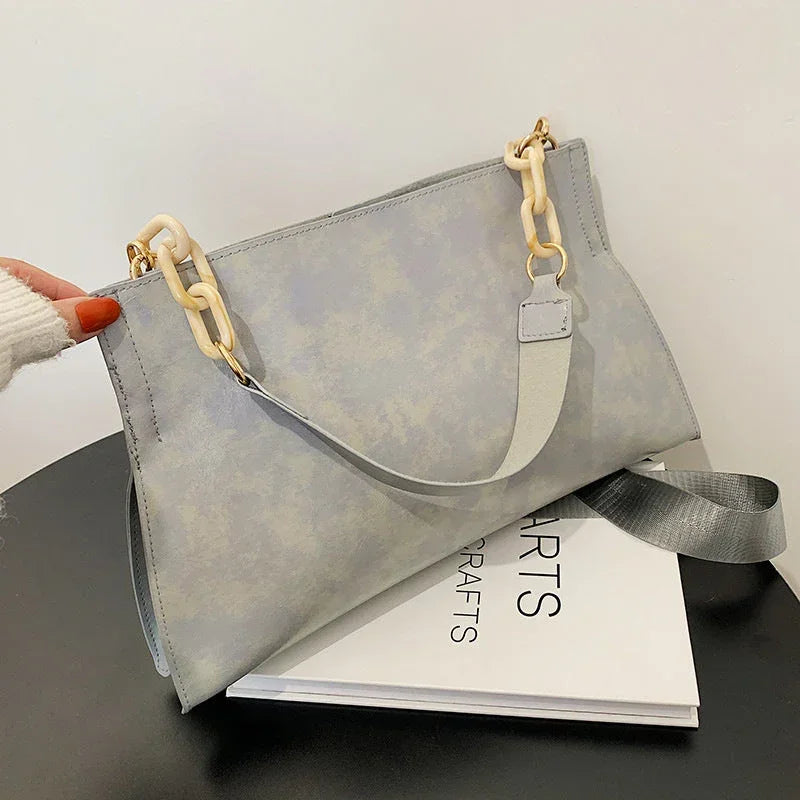 Light grey handbag with gold and cream chain strap on black table, minimalist style