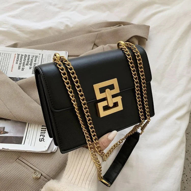 Black leather shoulder bag with gold chain strap and geometric clasp on beige coat and newspaper