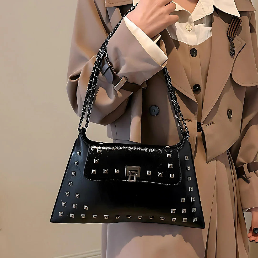 Woman in beige trench coat holding black studded leather shoulder bag with chain strap
