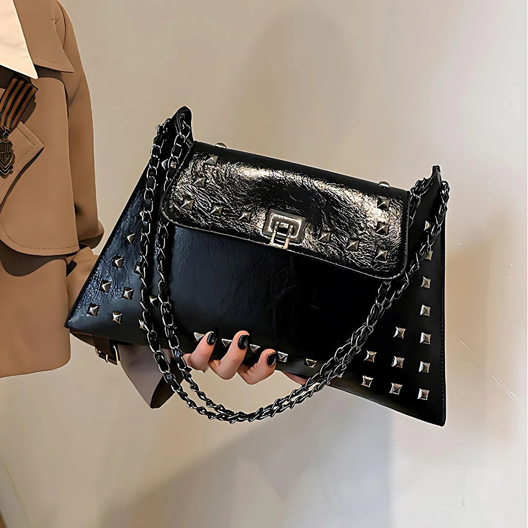 Glossy black studded handbag with chain strap and silver hardware, held by a woman in a tan coat
