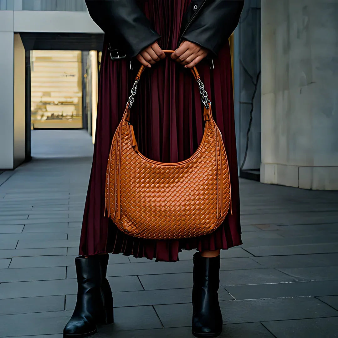 Woman holding woven brown leather hobo bag, wearing black boots and burgundy pleated skirt