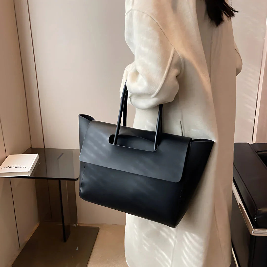 Woman in beige coat holding black minimalist leather tote bag indoors