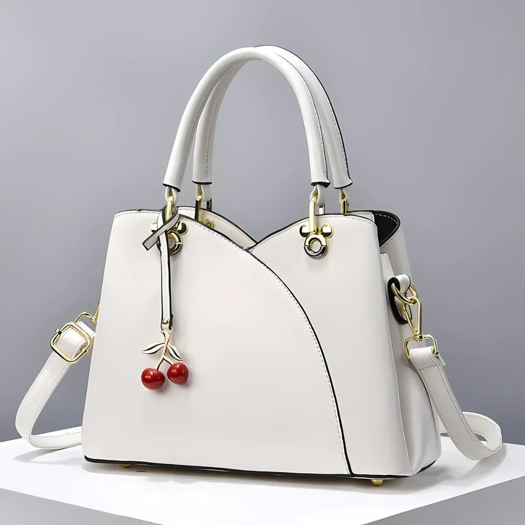 White leather handbag with gold hardware and cherry charm on neutral background