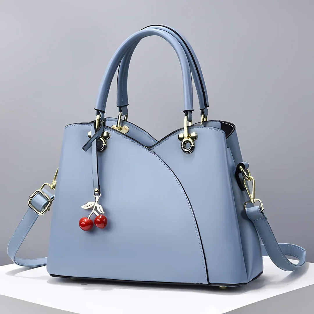 Light blue handbag with double handles, shoulder strap, and cherry charm on white display