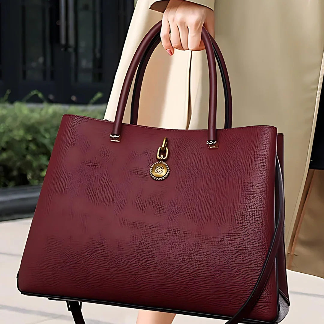 Woman holding large burgundy leather tote bag with gold charm, outdoor setting