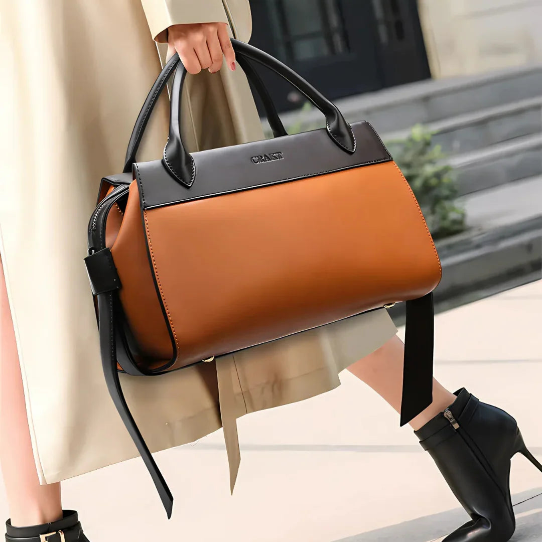 Woman holding large brown leather handbag with black handles, wearing trench coat and ankle boots