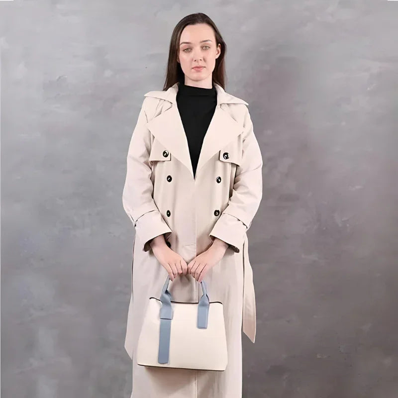 Woman in beige trench coat holding cream handbag with blue straps, gray background