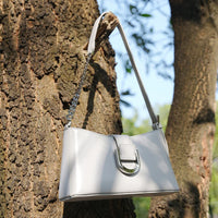 White leather shoulder bag with silver buckle detail hanging on a tree outdoors