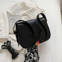 Black leather shoulder bag held by hand, on white textured jacket with book and accessory