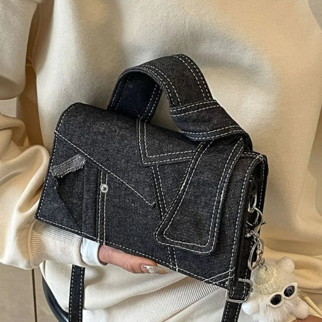 Black denim crossbody bag with white stitching and plush keychain, held by person in cream sweater