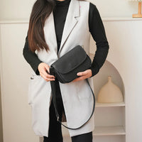 Woman in black top and gray vest holding a black leather crossbody bag indoors