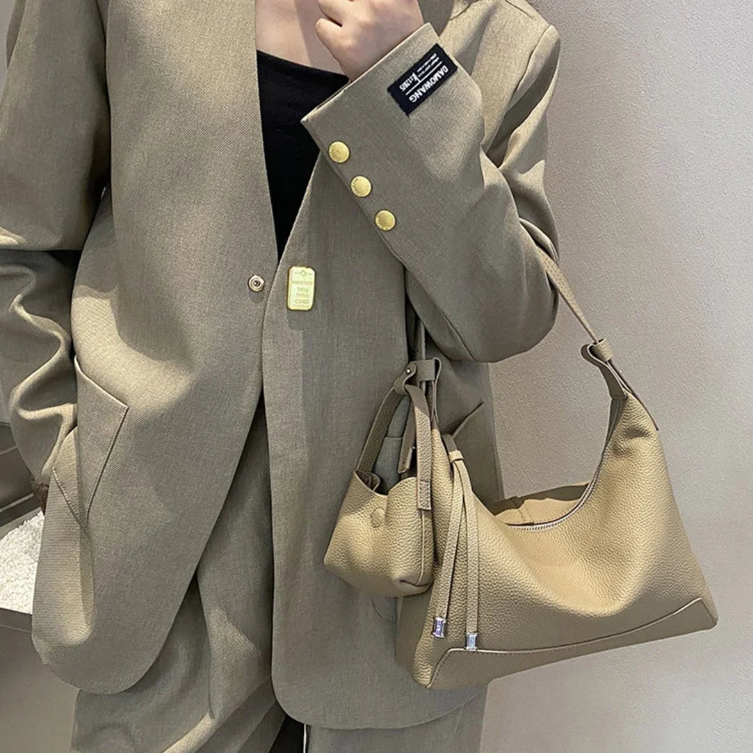 Woman in beige suit holding a tan leather shoulder bag against a neutral wall