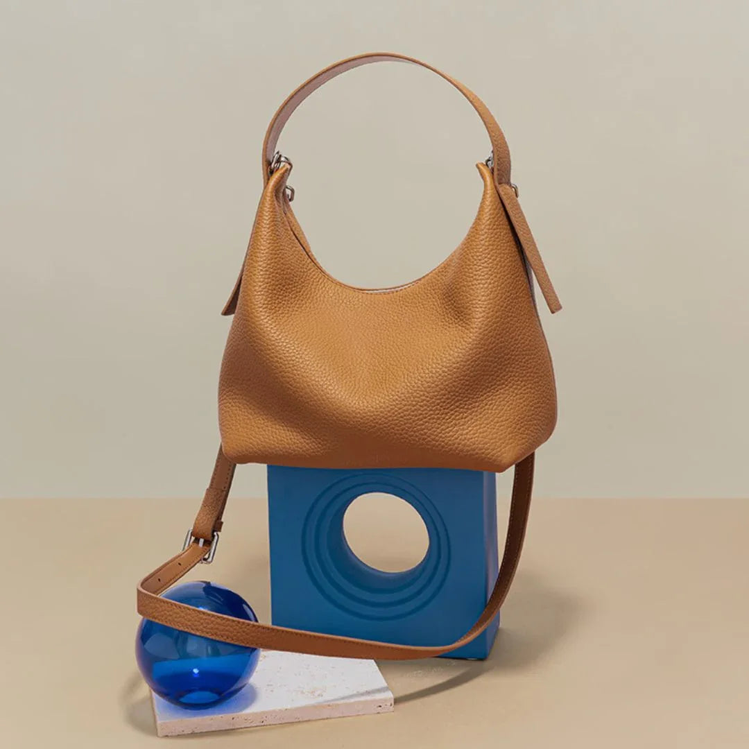 Tan leather handbag with shoulder strap, displayed on blue geometric stand and blue glass sphere