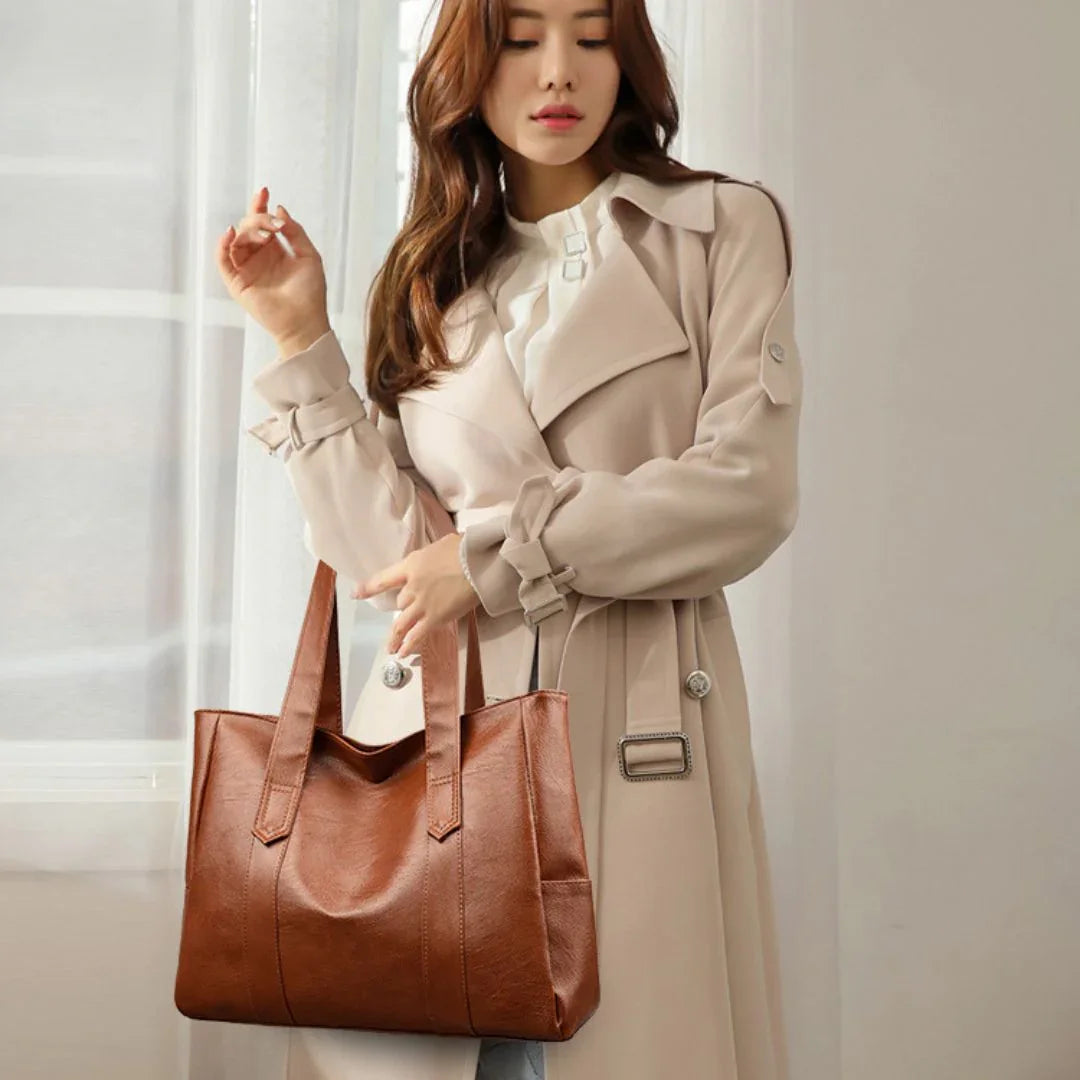 Woman in beige trench coat carrying large brown leather tote bag by window