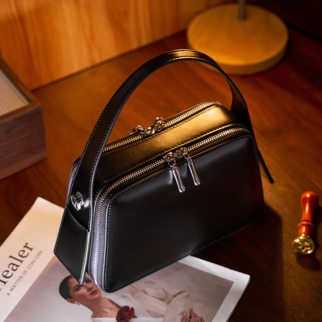 Black leather handbag with double zippers on a wooden desk beside a magazine and a lamp