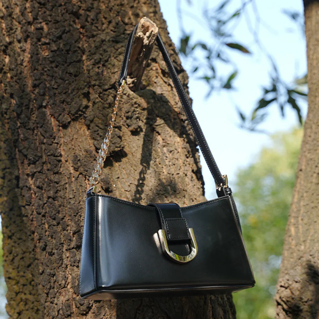 Black leather shoulder bag with gold chain and buckle detail hanging on a tree outdoors