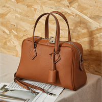 Brown leather handbag with handles and shoulder strap on beige table, wood panel background