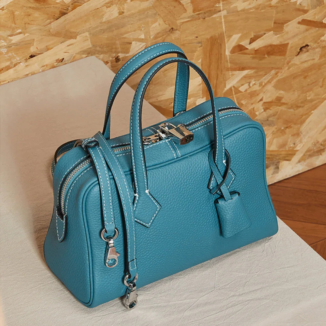 Teal leather handbag with silver hardware and lock on a light surface, wood background