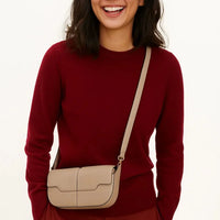Woman in red sweater wearing beige crossbody leather bag, smiling against plain background
